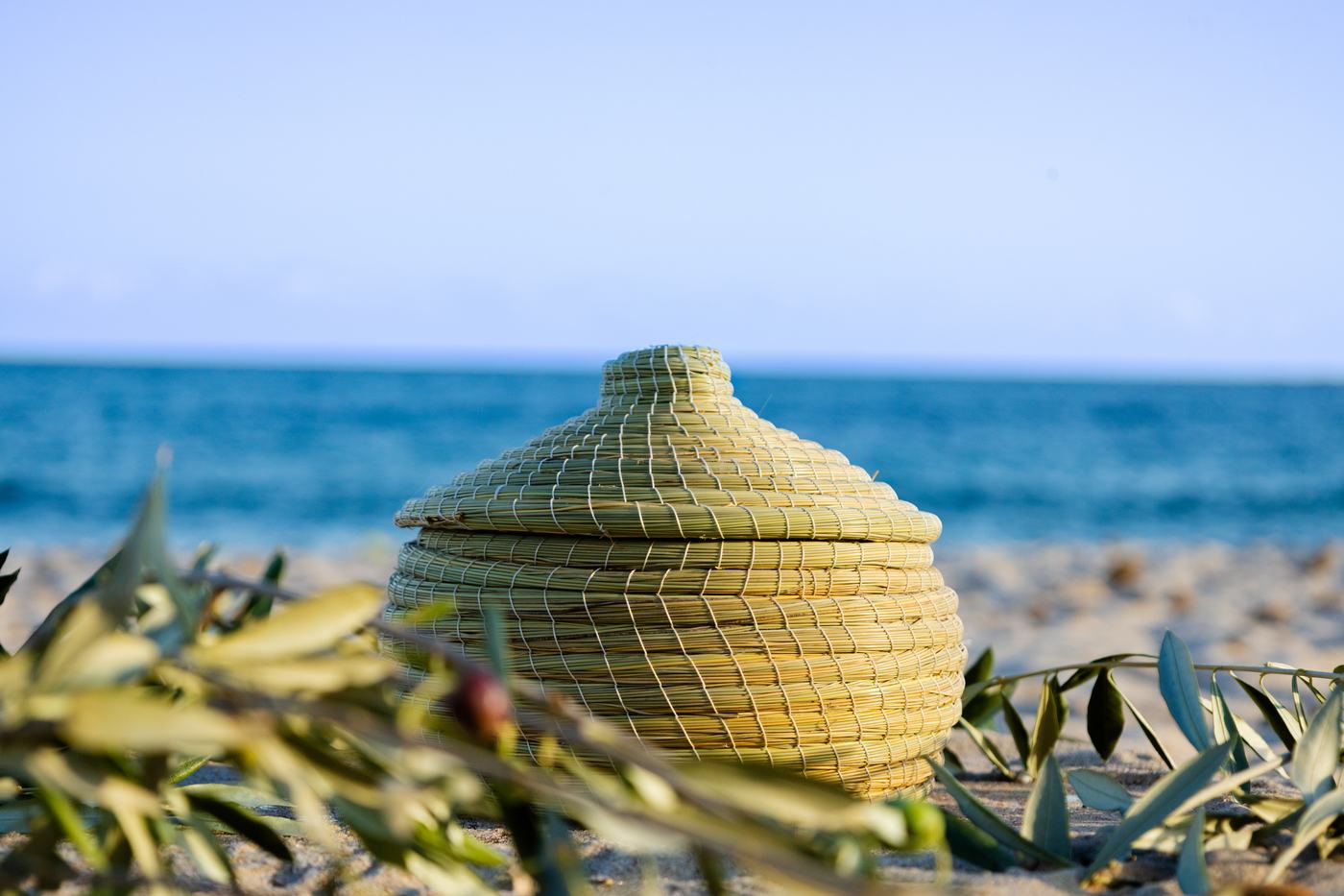 Un panier à couvercle en alfa posé dans le sable, entre des branches d’olivier