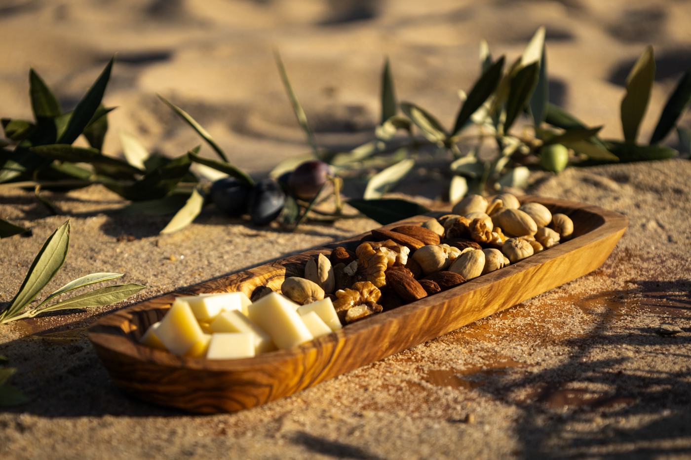 Un plateau allongé en bois d’olivier garni de fromages et de noix, dans le sable